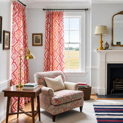 Red pink and orange geometric pinch pleat curtains in a bright living room with a fireplace, patterned armchair, brass lamp and classic decor